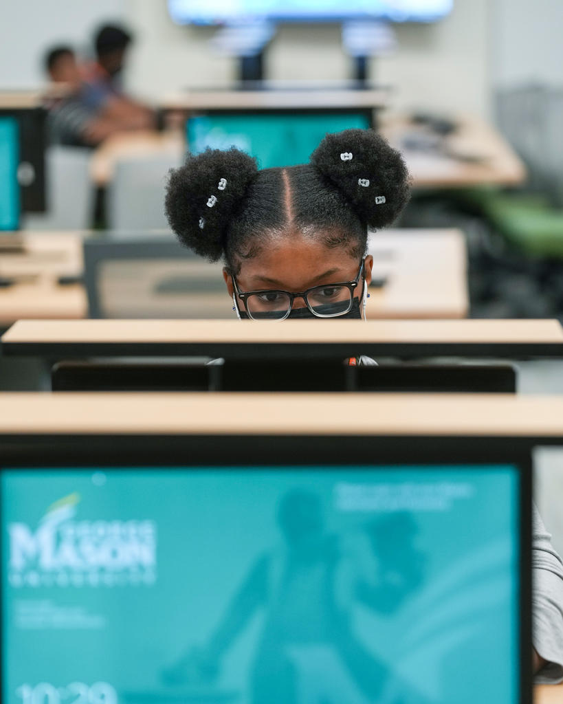 A student works on a computer before her. In the foreground of the shot is an out of focus computer monitor where the George Mason University logo can just be made out.
