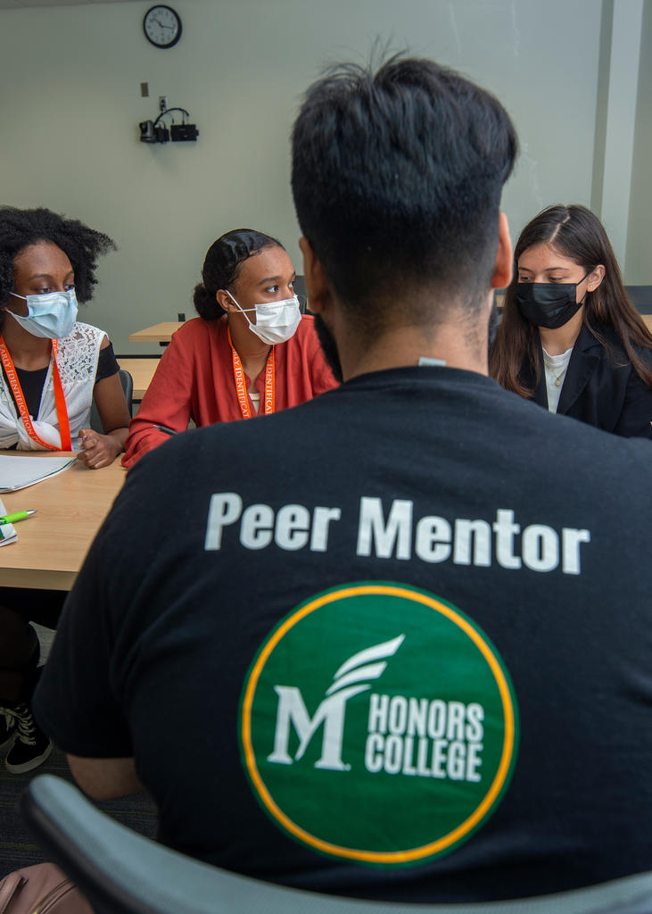 A student from the Mason Honors College in a Peer Mentor shirt faces a group of students, presumably talking with them.
