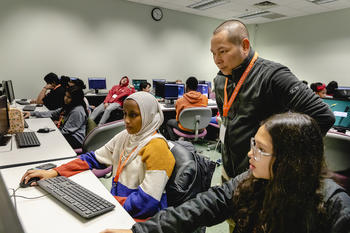 A tutor stands over two students in a crowded computer lab, providing guidance